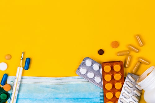 Protective medical mask on yellow background surrounded with colorful pills. Creative concept of healthcare, hygiene, virus protection and medical treatment on coronavirus quarantine