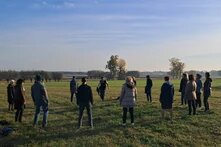 Photo: A group of people stand in a circle in a meadow. Fields, trees and a clear sky can be seen in the background.
