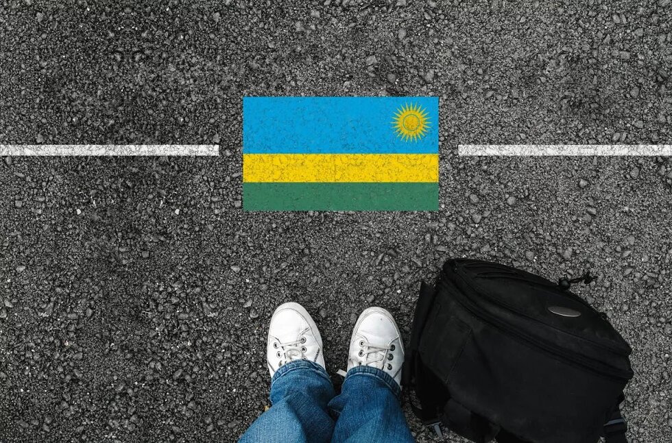 A man with a shoes and backpack is standing on asphalt next to flag of Rwanda and border